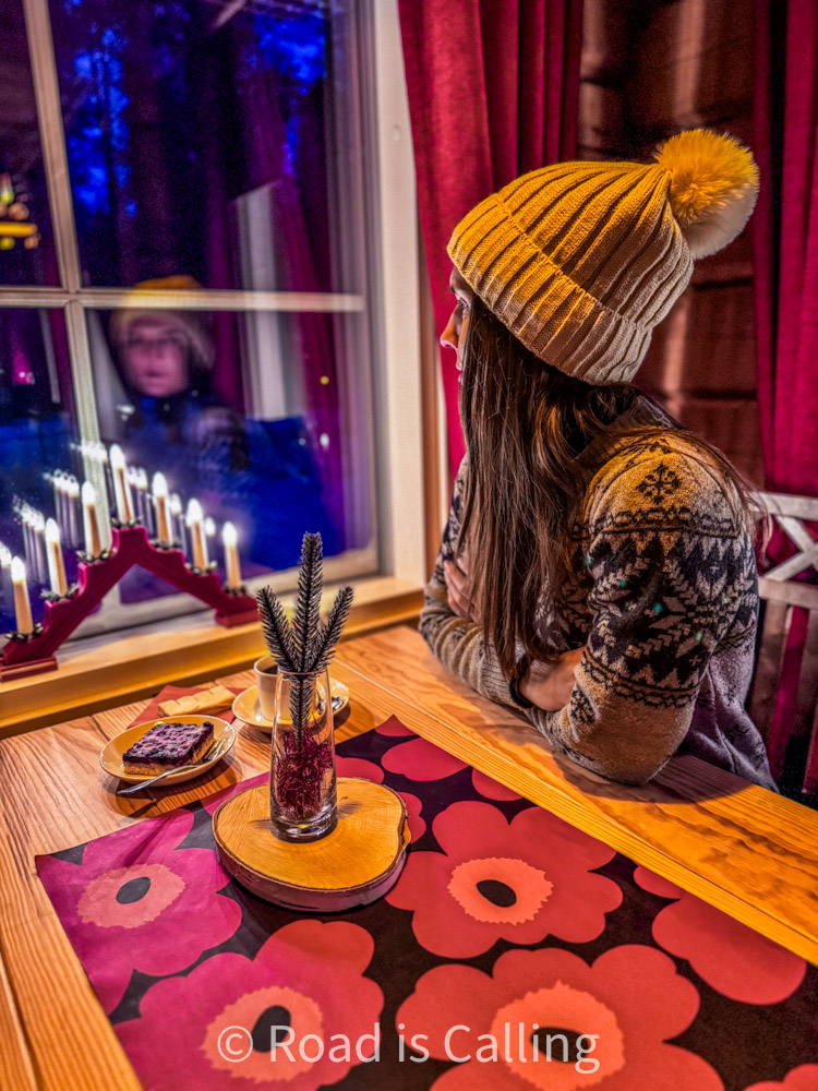 Woman in wool hat sitting by window with candle and cake in cozy Lapland cafe