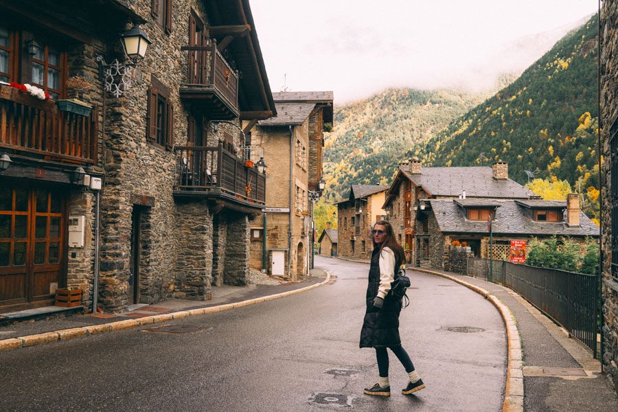 Woman wearing a long black coat and sneakers walking along a quiet, winding street in a picturesque mountain village with stone buildings and wooden balconies, surrounded by green and autumn-colored trees under a cloudy sky.