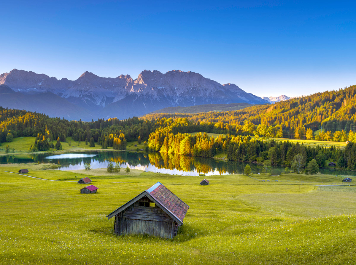 view of the alpine meadow and wooden huts with a lake and mountain backdrop by Geroldsee