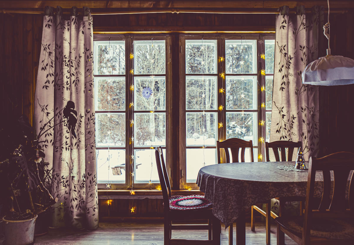 living room with a table, flower, lamp and wide windows in a forest cabin in Estonia with snowy view outside