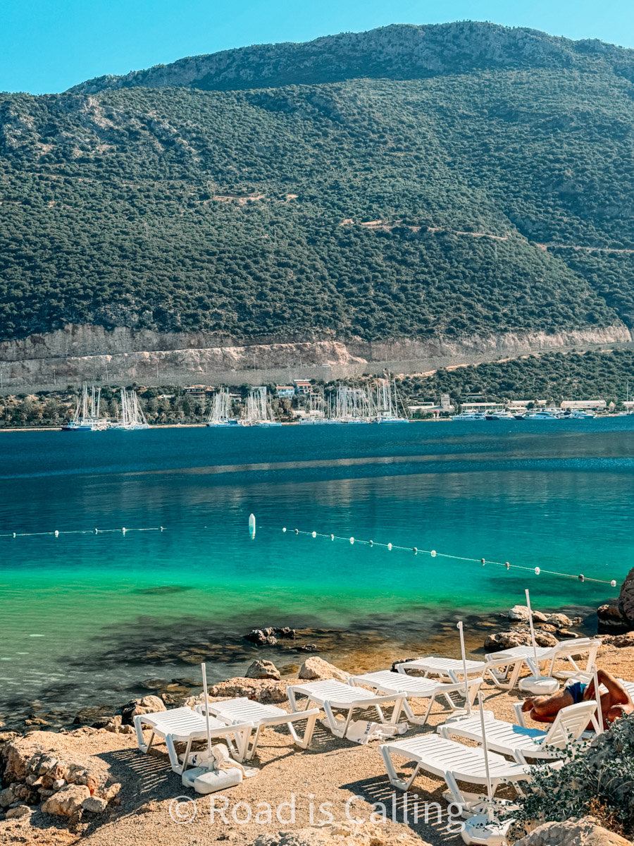 Peaceful beach in Kaş, Turkey with white sun loungers, emerald-green water, and mountain views across the bay