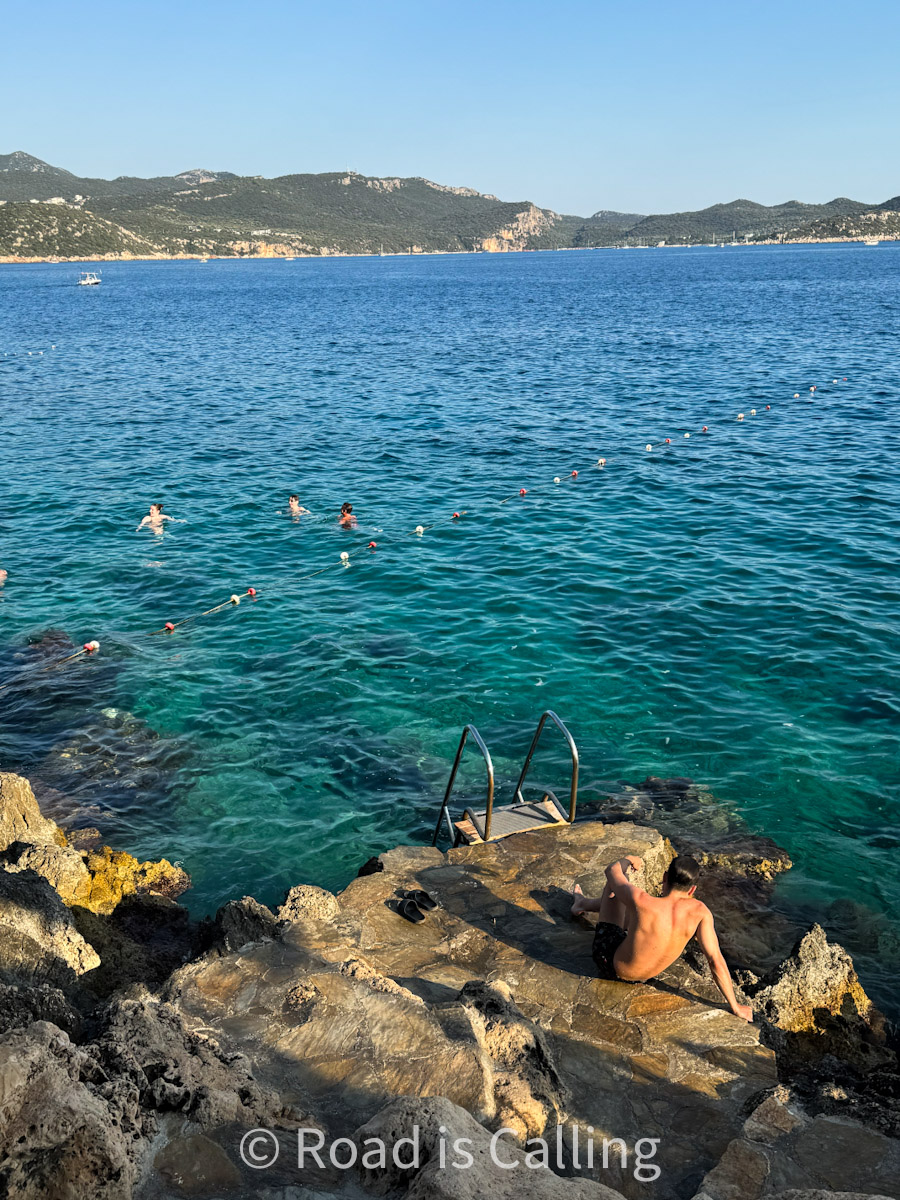 Rocky beach platform in Kaş, Turkey with a metal ladder into the turquoise sea and people swimming in the background