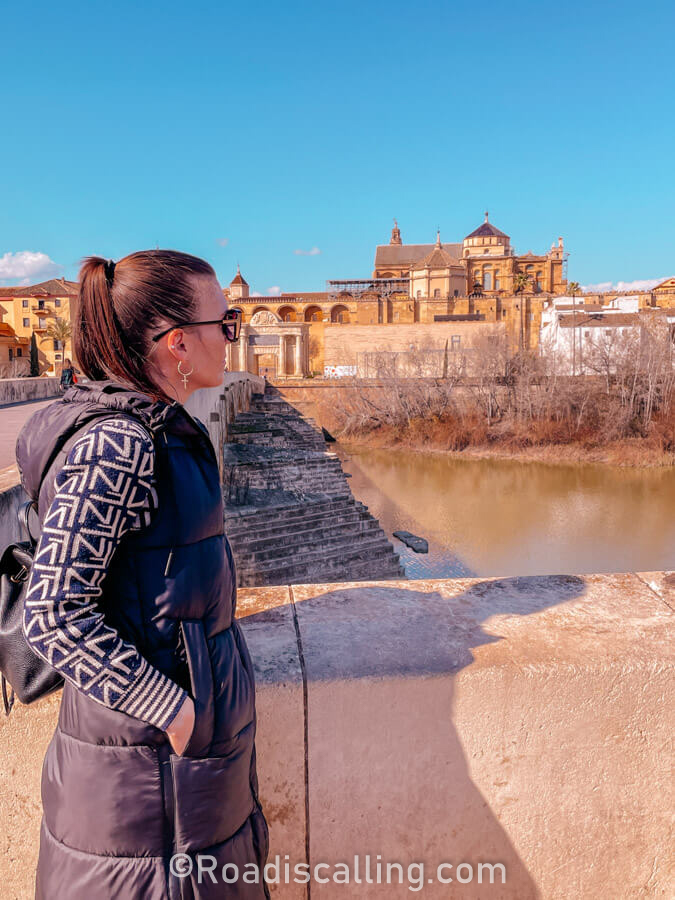 woman standing on a historic bridge in Cordoba in winter, overlooking the river and cathedral