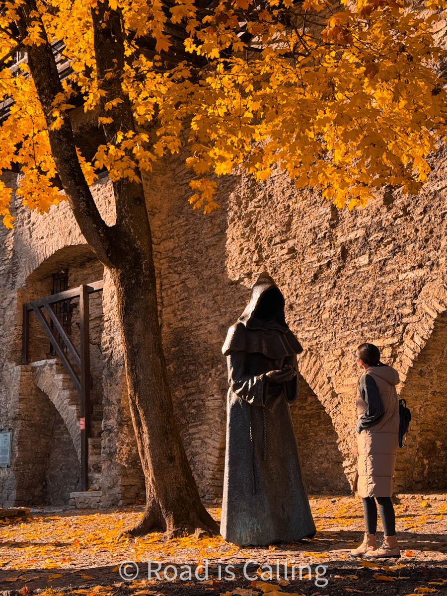 Woman standing under golden autumn leaves beside a cloaked monk statue in Tallinn’s medieval walls